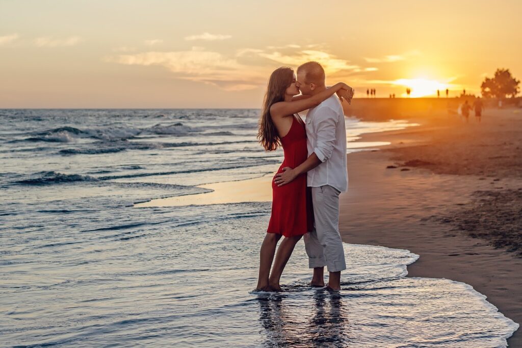 A man and woman embrace on the beach, silhouetted against a vibrant sunset sky.