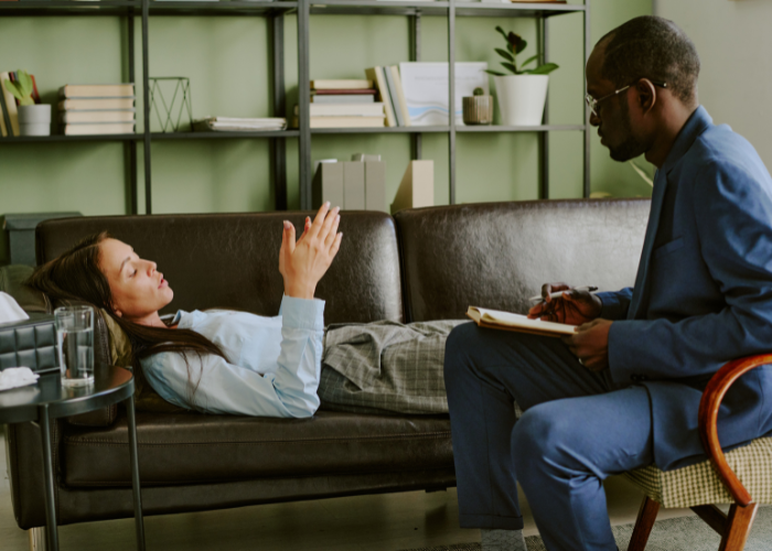 A man and woman sit on a couch, engaged in a conversation, smiling and appearing relaxed.
