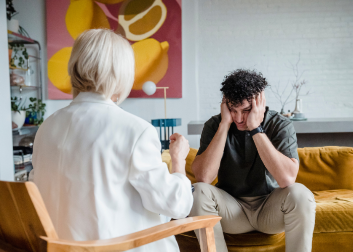 A man sits on a couch with his head in his hands, while a woman sits beside him, looking concerned.